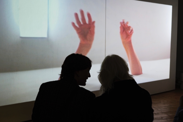 Photo of back of two people sitting watching a large screen.  The screen has two huge human hands projected onto it. 